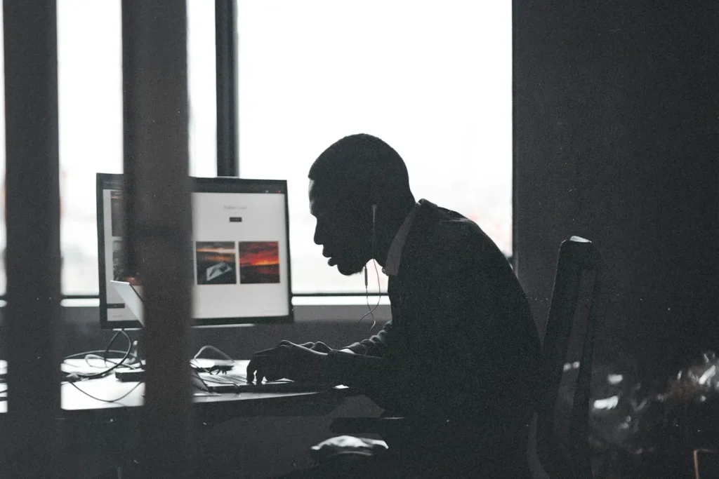 A person sits at a desk working on a computer with large windows behind them, silhouetted against the light