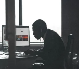 A person sits at a desk working on a computer with large windows behind them, silhouetted against the light