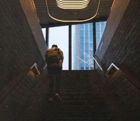 A person with a backpack walks up a dimly lit stairway toward large windows overlooking tall modern buildings outside