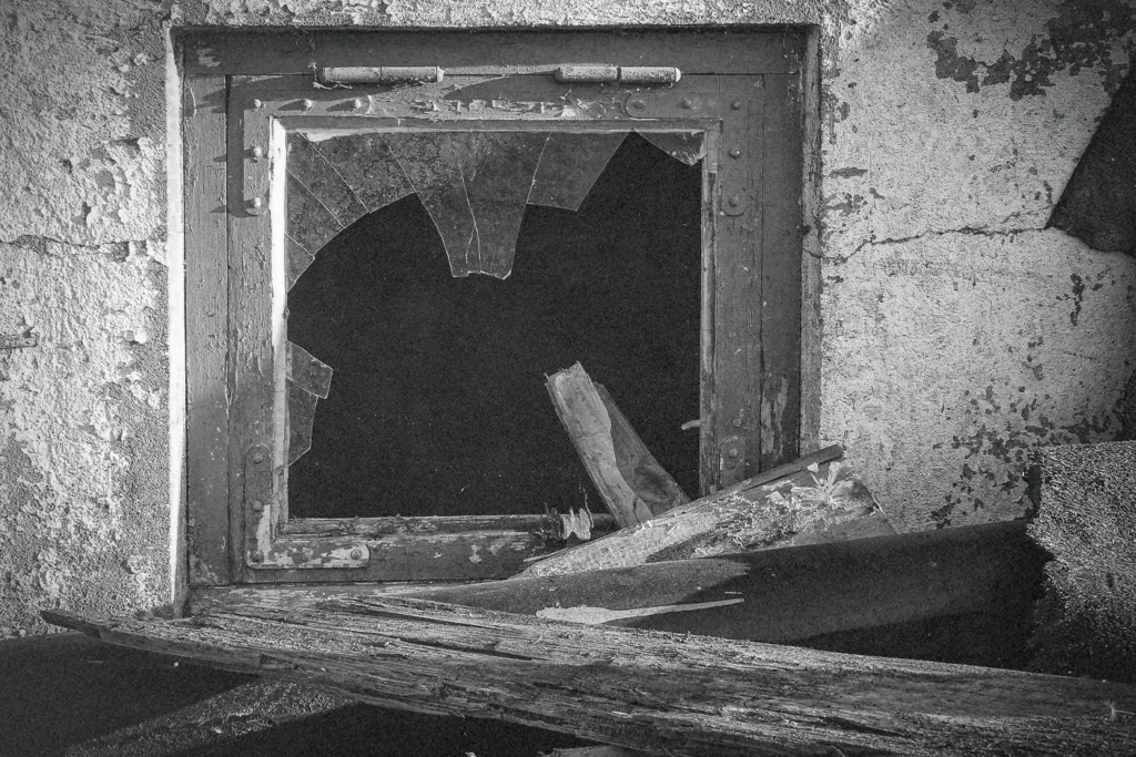 A broken window with shattered glass sits in a weathered concrete wall surrounded by debris