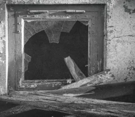 A broken window with shattered glass sits in a weathered concrete wall surrounded by debris