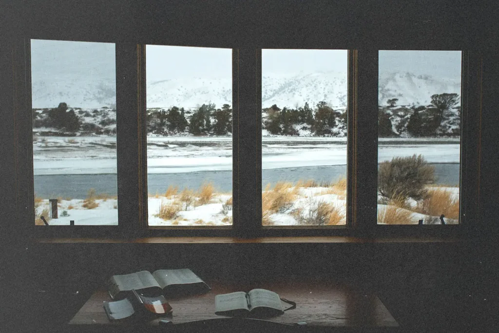 Open Bibles on a wooden table inside a cabin, overlooking a snowy landscape and frozen river through large windows