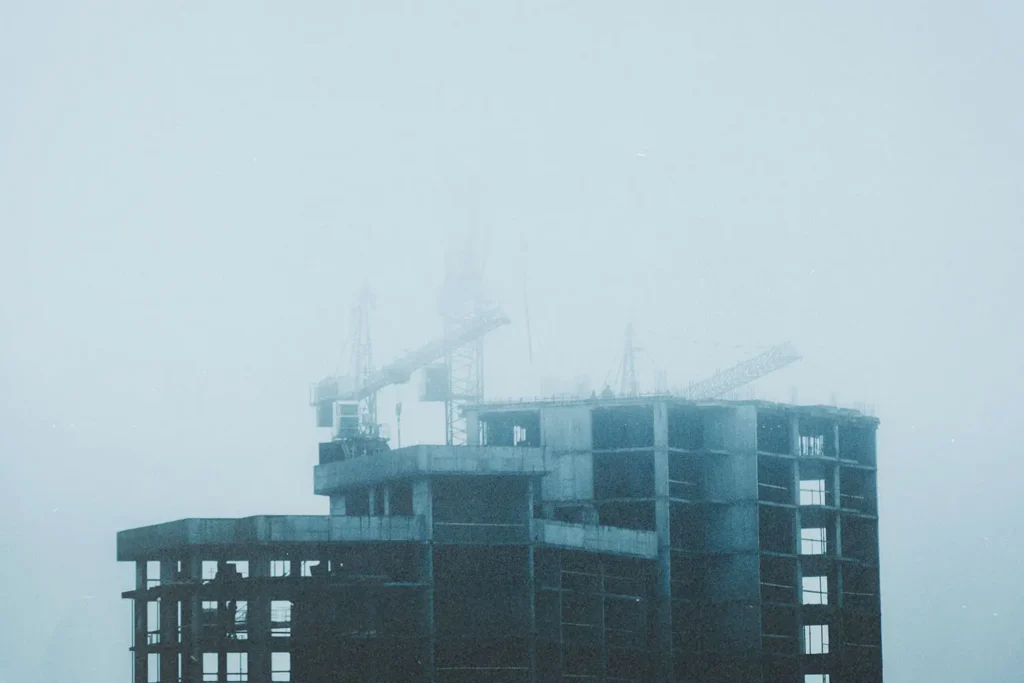 A tall building under construction stands in thick fog, with cranes barely visible through the haze