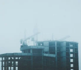 A tall building under construction stands in thick fog, with cranes barely visible through the haze