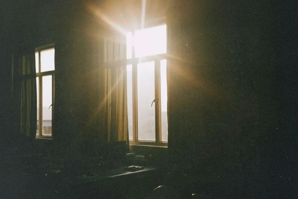 Sunlight streams through tall window panes, casting bright rays and soft golden light across a dimly lit room