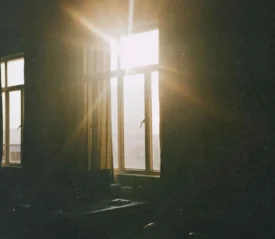 Sunlight streams through tall window panes, casting bright rays and soft golden light across a dimly lit room