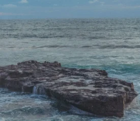 A large flat rock formation sits in the ocean as waves wash gently over its edges under a soft blue sky