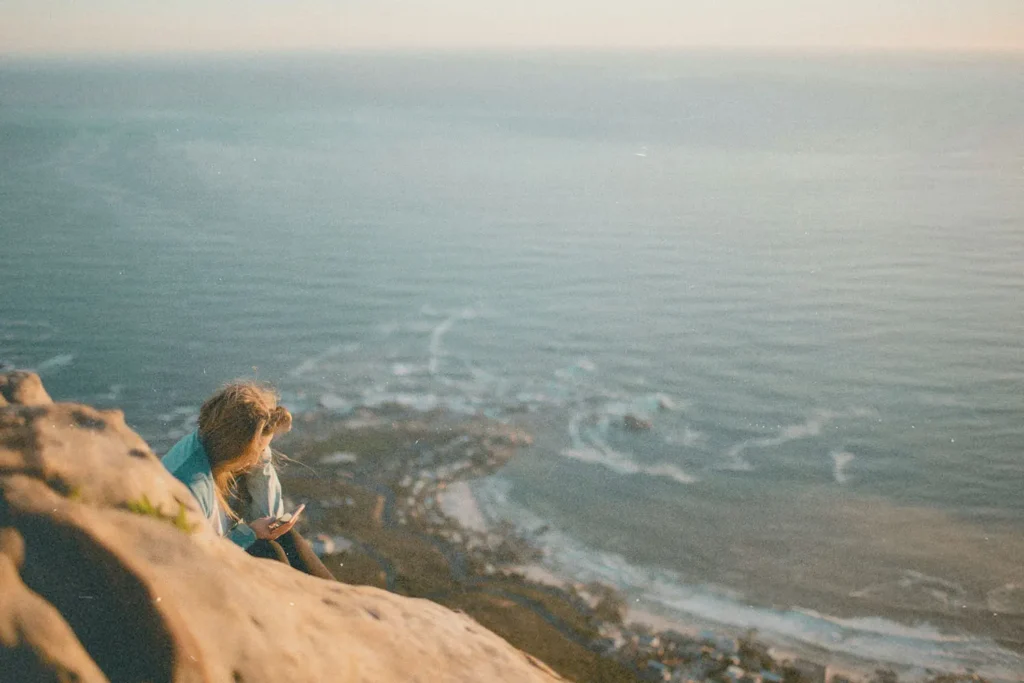 Person sitting on a rocky cliff overlooking the ocean at sunset, with waves and coastline visible far below