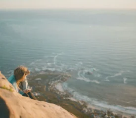 Person sitting on a rocky cliff overlooking the ocean at sunset, with waves and coastline visible far below