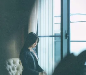 Person sitting in a softly lit room, looking out a large window with sheer curtains and an urban view outside