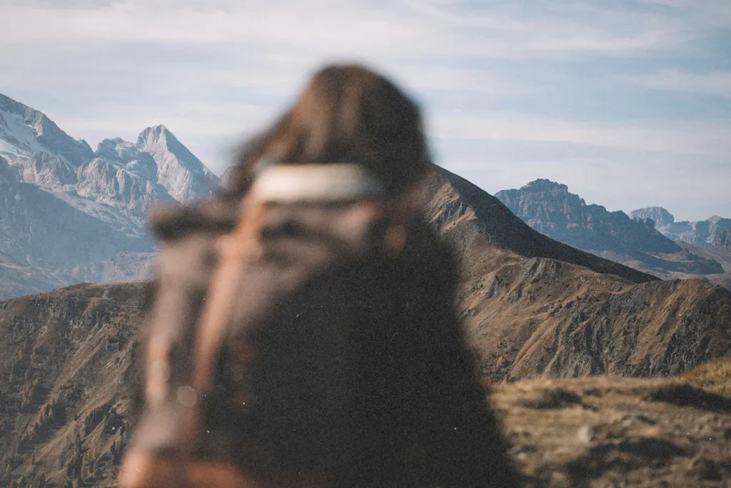A person with a backpack stands on a mountain trail, facing a distant range of rugged peaks
