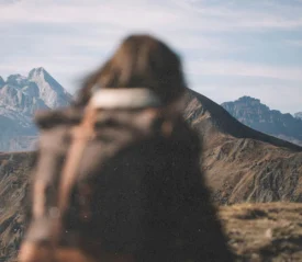 A person with a backpack stands on a mountain trail, facing a distant range of rugged peaks
