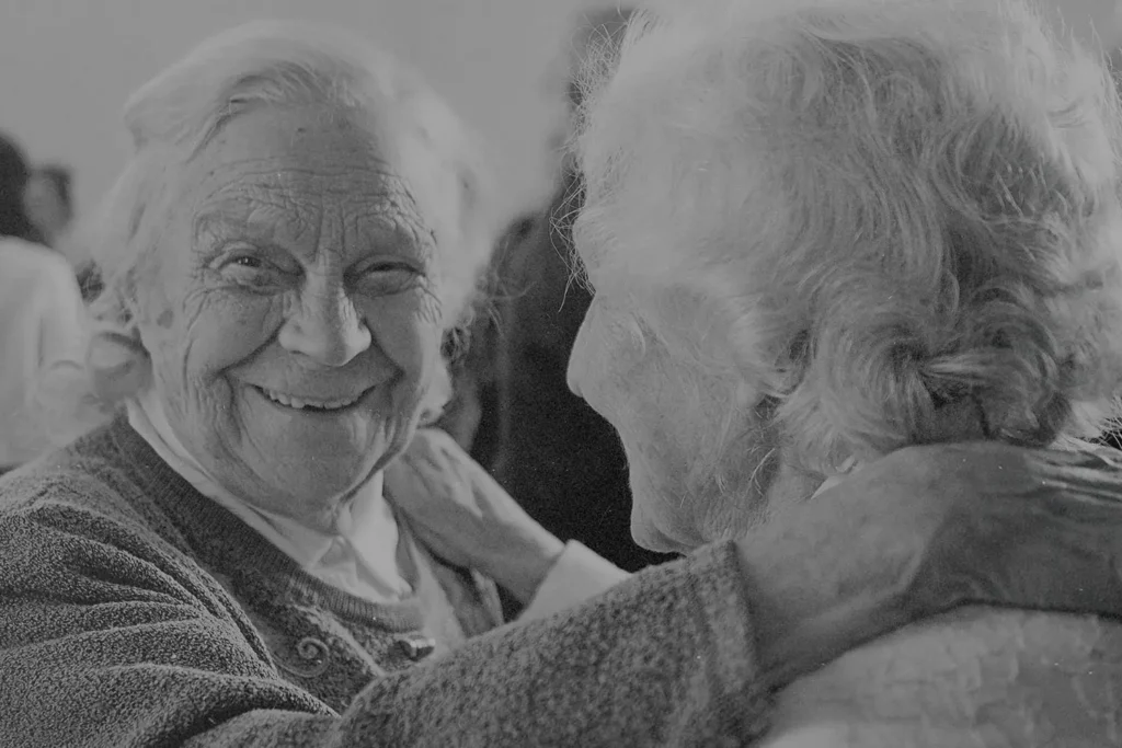 Two older adults embrace closely, one placing a hand gently on the other’s shoulder