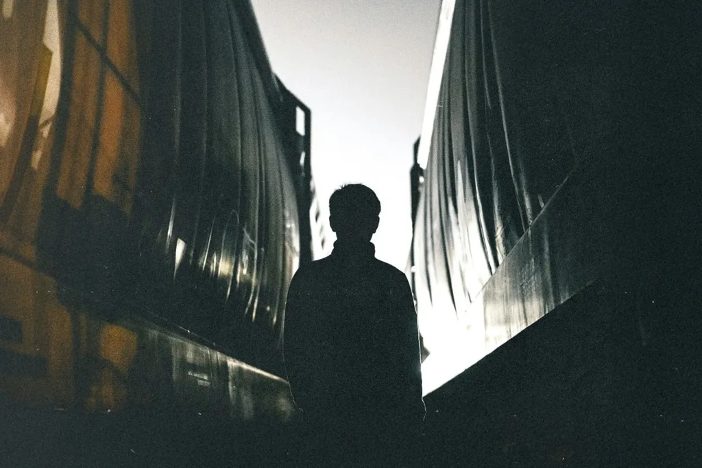 A silhouette of a person stands between two train cars in low light, with bright illumination behind them