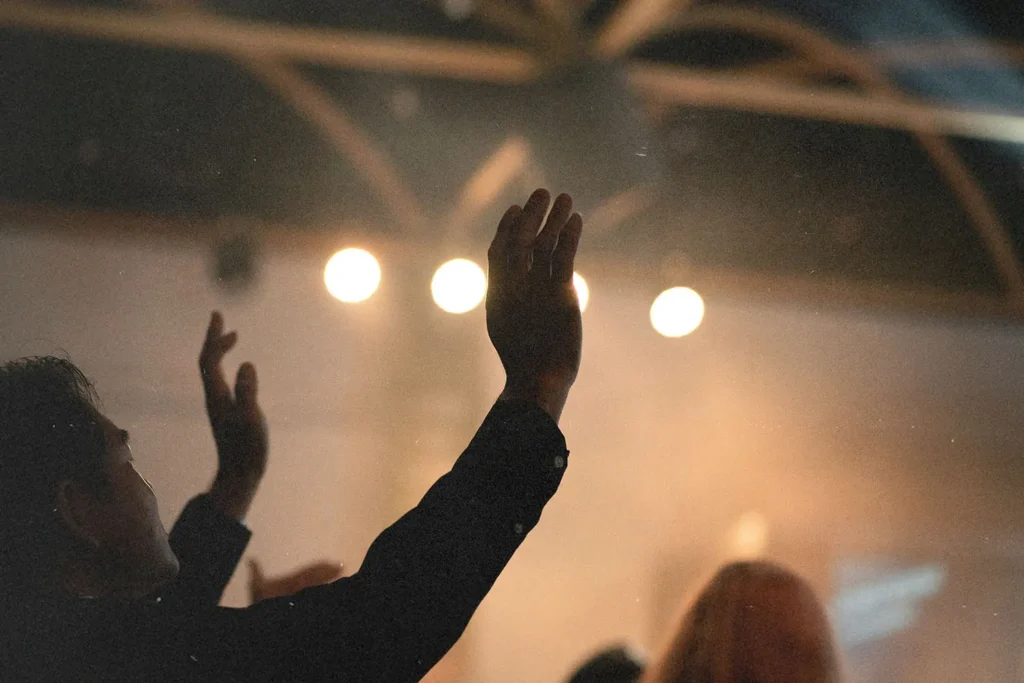 People raise their hands in worship inside a dimly lit room with bright stage lights overhead
