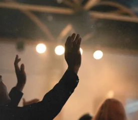 People raise their hands in worship inside a dimly lit room with bright stage lights overhead