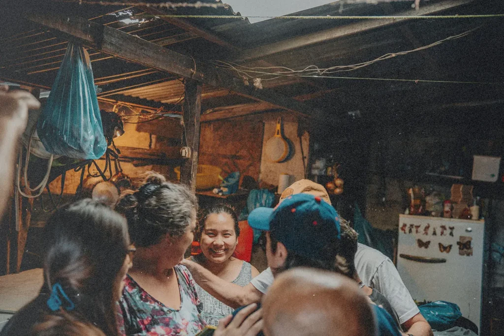 Several people gather around someone in a rustic, dimly lit shelter, placing comforting hands on their shoulders