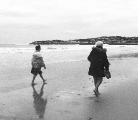 Two people walking barefoot along a quiet beach shoreline on a cloudy day, with a town visible in the distance