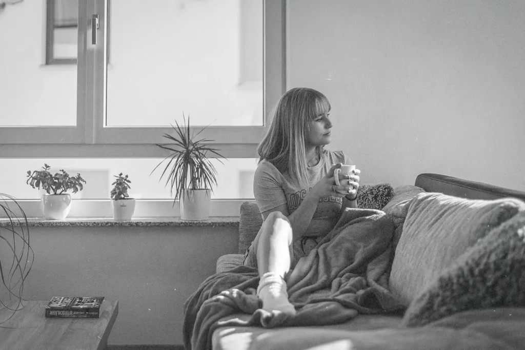 Person relaxing on a sofa with a blanket and holding a warm drink near a bright window with potted plants