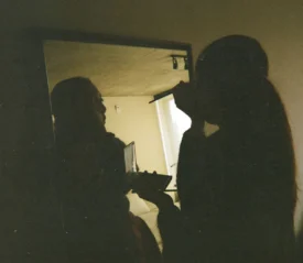 Person applying makeup in front of a mirror in a dimly lit room, holding a compact and brush