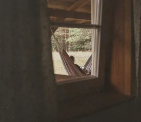 View through a window of a person reclining in a striped hammock under a wooden porch, with trees and grass in the background