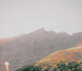 Rugged mountain landscape with steep rocky slopes rises beneath a hazy sky, with autumn-colored grass and scattered trees in the foreground