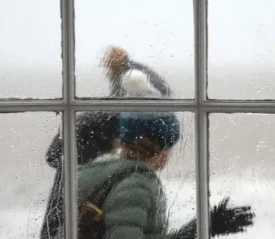 Two people in winter clothing walk past a rain‑covered window, their figures softened by the water droplets on the glass