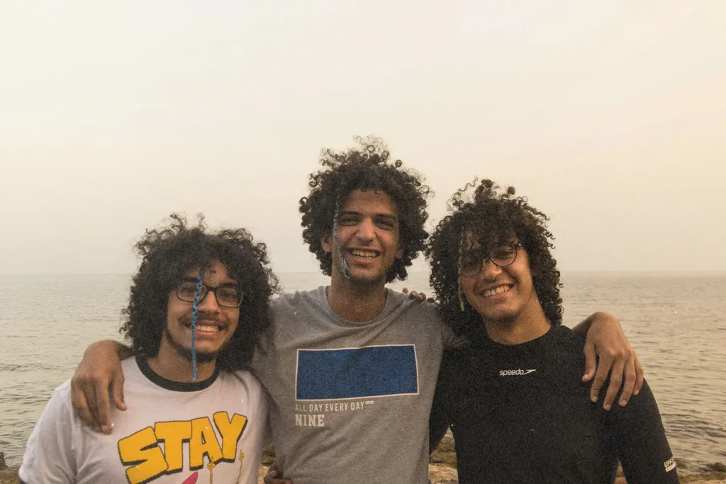 Three friends stand arm in arm near the shoreline, with the sea and sky forming a soft backdrop
