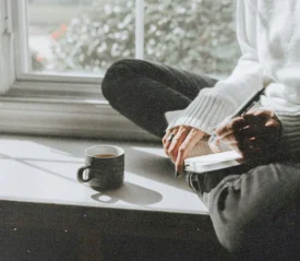 Person sitting on a sunny windowsill with a notebook and a cup of coffee, writing in soft natural light
