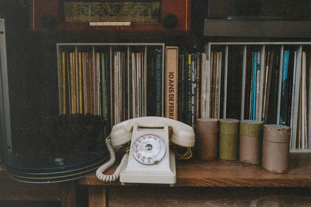 Vintage rotary telephone displayed on a wooden shelf surrounded by vinyl records, books, and small storage canisters