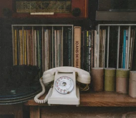 Vintage rotary telephone displayed on a wooden shelf surrounded by vinyl records, books, and small storage canisters