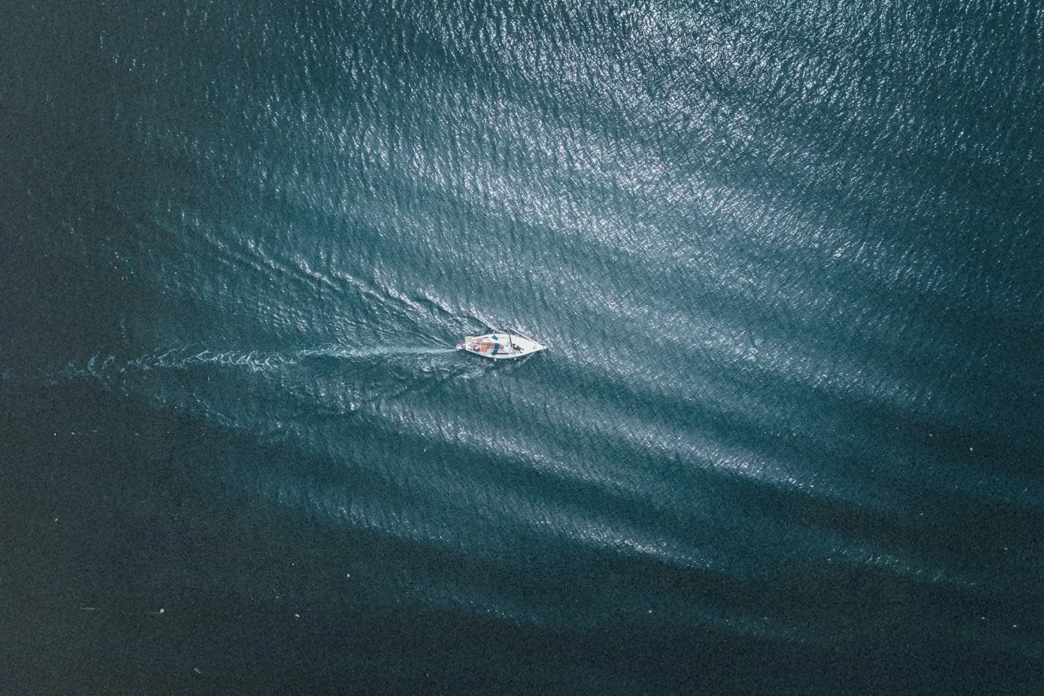 “Aerial view of a small boat moving across deep blue water, leaving a soft wake behind it