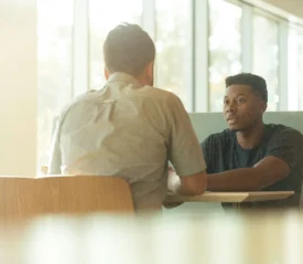 Two people sitting across from each other at a table in a bright room with large windows, engaged in conversation