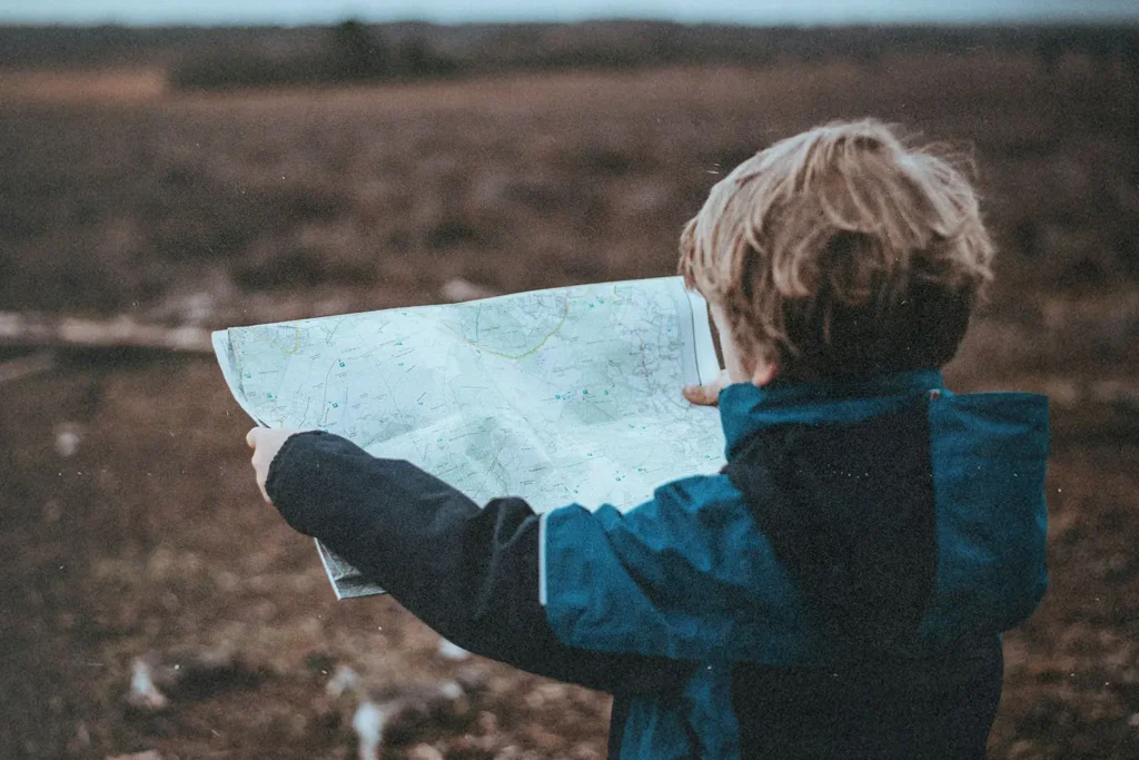 A child wearing a blue jacket stands outdoors holding a large map, looking out over a wide, open landscape