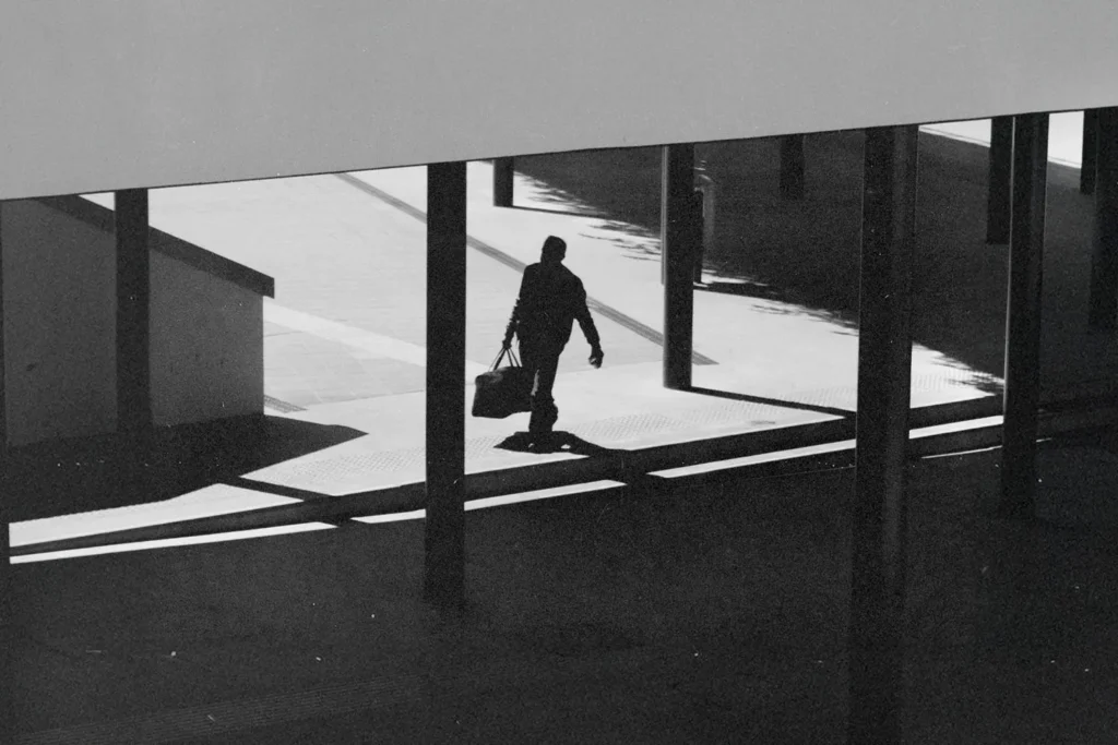 A silhouetted figure walks through a modern outdoor walkway carrying a bag, with strong shadows cast across the ground