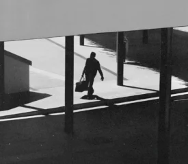A silhouetted figure walks through a modern outdoor walkway carrying a bag, with strong shadows cast across the ground