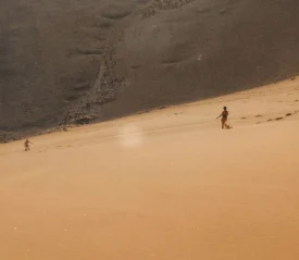 Two people walk across a vast desert slope, appearing small against the large sandy landscape