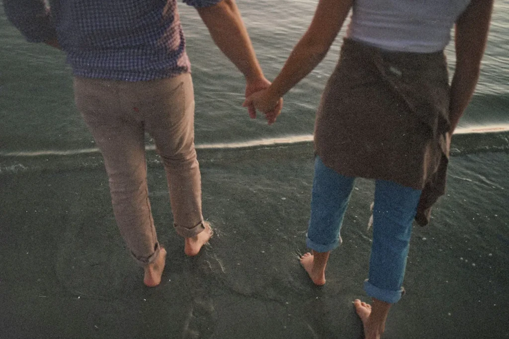 Two people holding hands while walking barefoot along the shoreline where the water meets the sand