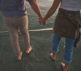 Two people holding hands while walking barefoot along the shoreline where the water meets the sand
