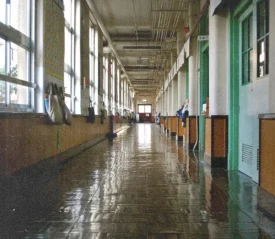 A long, empty school hallway lined with windows and classroom doors reflects bright light on its shiny floor