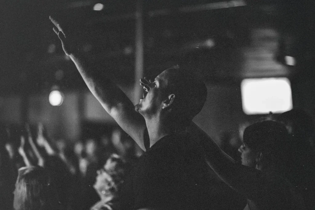 Person raising an arm in worship during a dimly lit church or concert gathering with others in the background