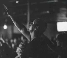 Person raising an arm in worship during a dimly lit church or concert gathering with others in the background