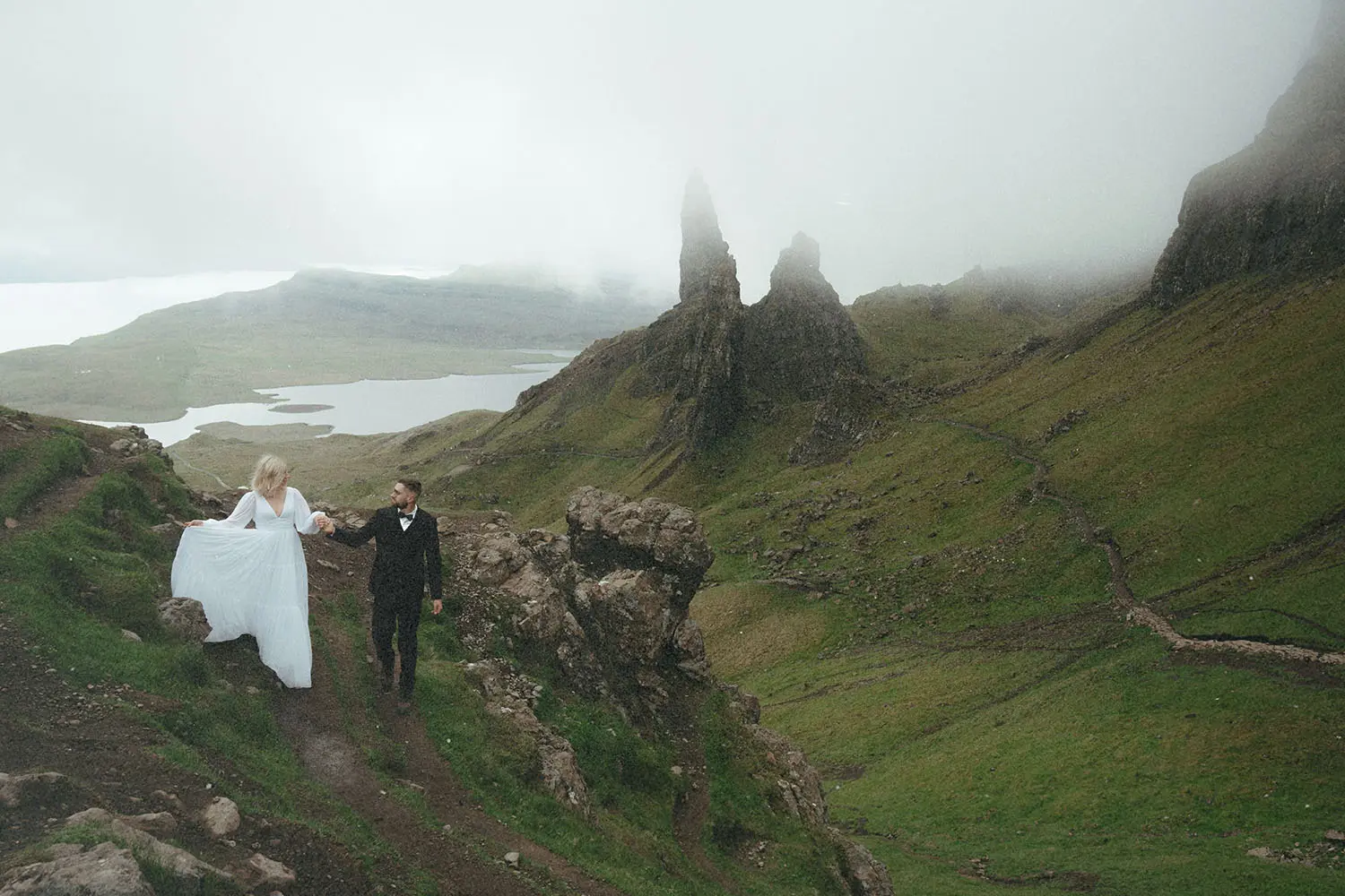 ride and groom holding hands while hiking through mist-covered highlands with steep cliffs and rolling green hills