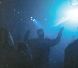 A silhouetted crowd stands in blue stage lighting, with one person raising both hands toward a bright spotlight