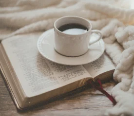 White coffee cup resting on an open Bible surrounded by a soft, cozy blanket on a wooden surface