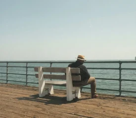 A person wearing a hat sits alone on a wooden pier bench, looking out over pier railing at a calm ocean