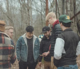 Group of people gathered in a wooded outdoor area, standing together among trees and fallen leaves