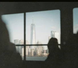 City skyline framed through a window, featuring a tall modern skyscraper and surrounding buildings with silhouetted observers in the foreground