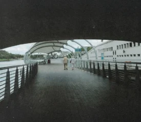 Two people walk together along a covered riverside boardwalk with railings on both sides