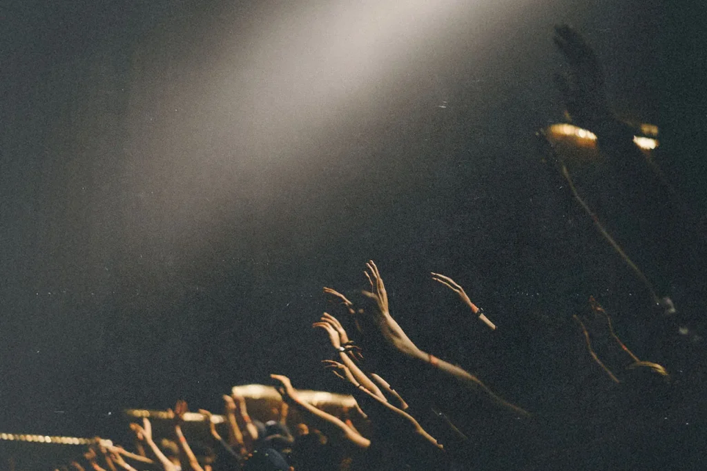 Dozens of raised hands reach toward a bright spotlight in a dimly lit gathering